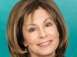 Woman with shoulder-length light brown hair smiling, wearing a blue jacket in front of a teal background