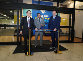 Three men dressed in suits posing with large scissors cutting a blue ribbon in front of an entrance to a physical therapy facility